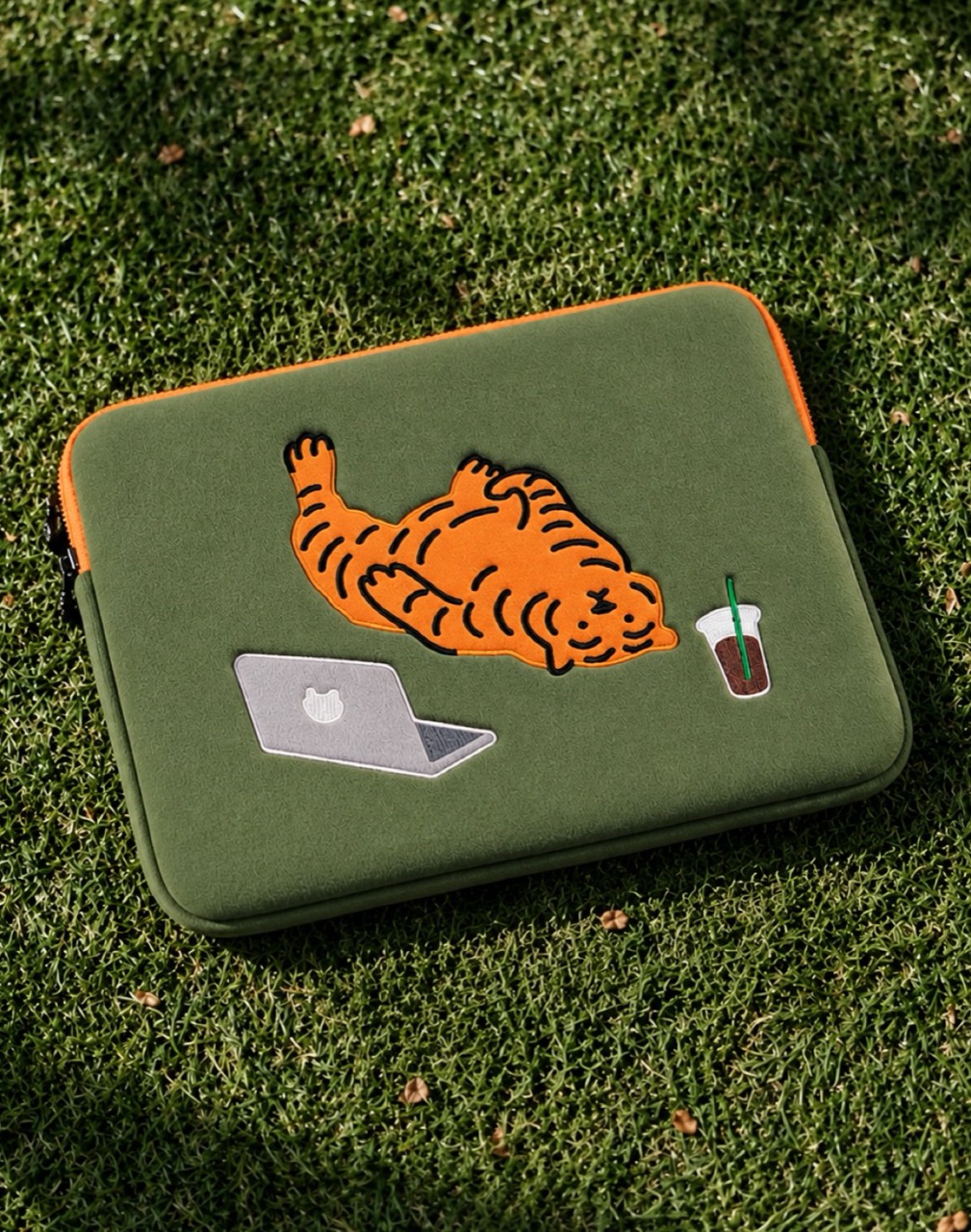 Green laptop pouch on grass, decorated with an orange tiger lying beside a laptop and an iced drink.